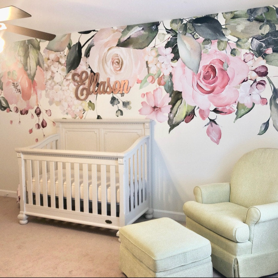 A styled nursery room featuring a large watercolor floral wall mural with pink roses and green leaves, a white wooden crib, a light green armchair, and an ottoman. A wooden name sign reading 'Ellason' is mounted on the wall.